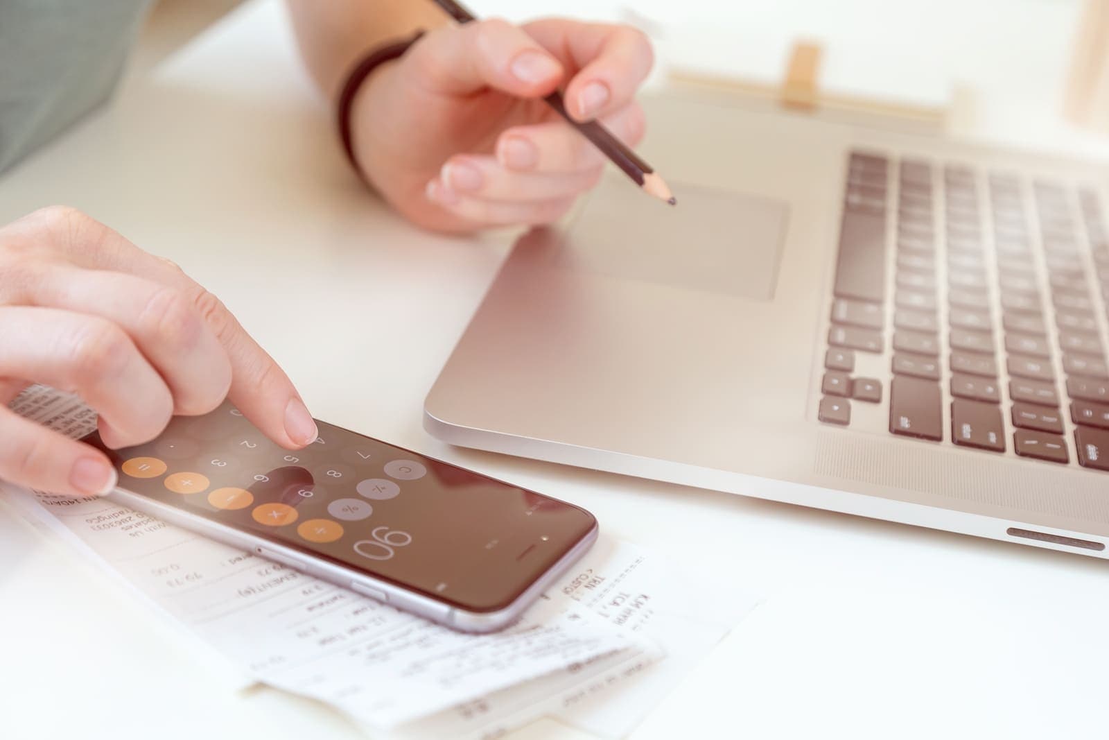 Hands using a phone calculator beside receipts and a laptop